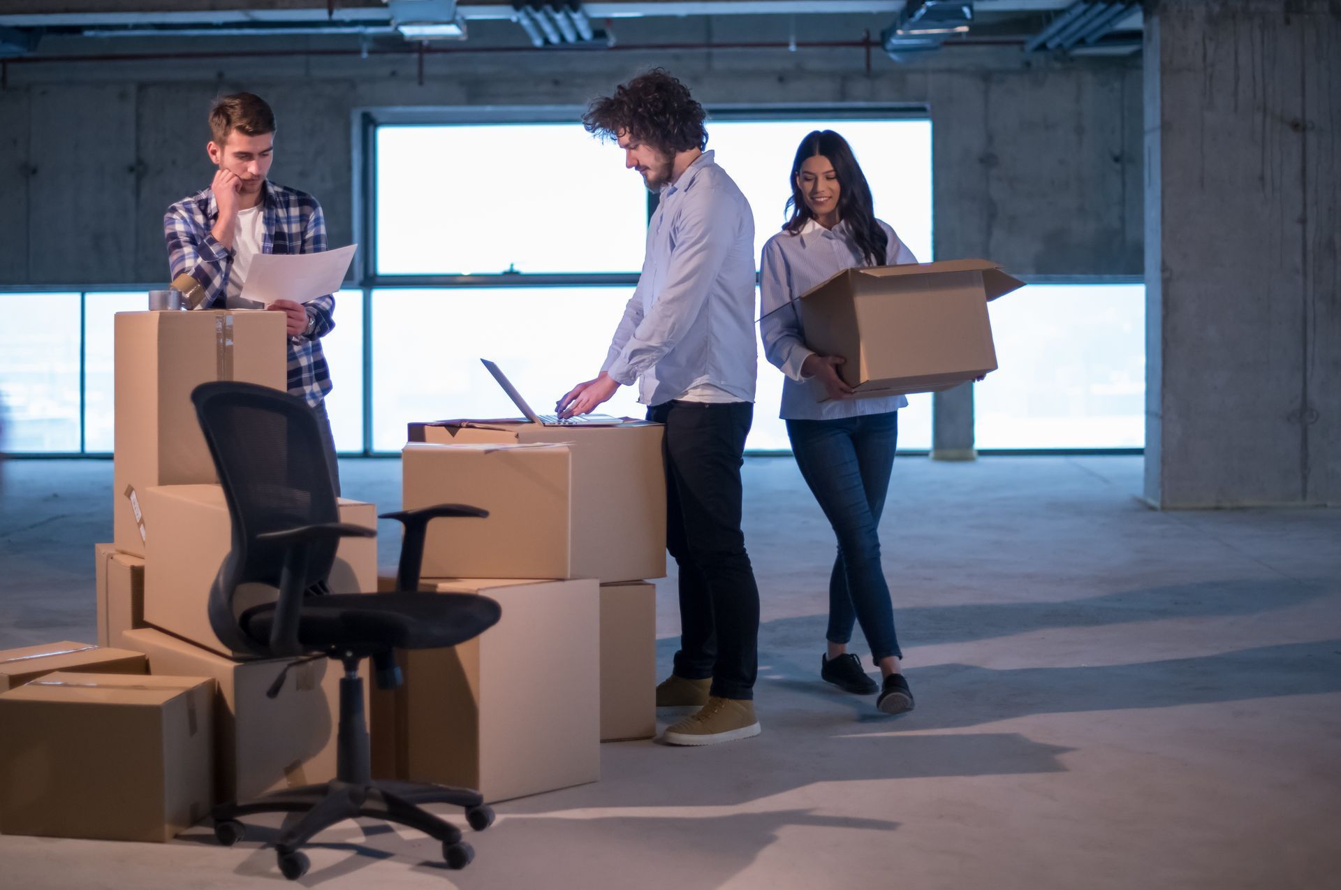 People moving boxes in an empty office with chairs and a desk, suggesting a relocation or move.