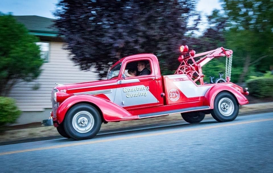 Red vintage tow truck driving on a road, trees and building in the background. Driver visible.