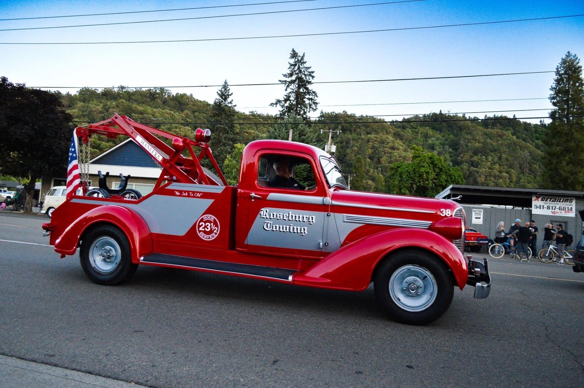 Red and grey vintage tow truck driving on a road. American flag on the back.