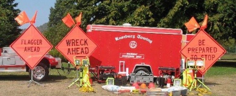Orange road signs warn of a flagger, wreck, and to be prepared to stop. A red trailer is in the background.