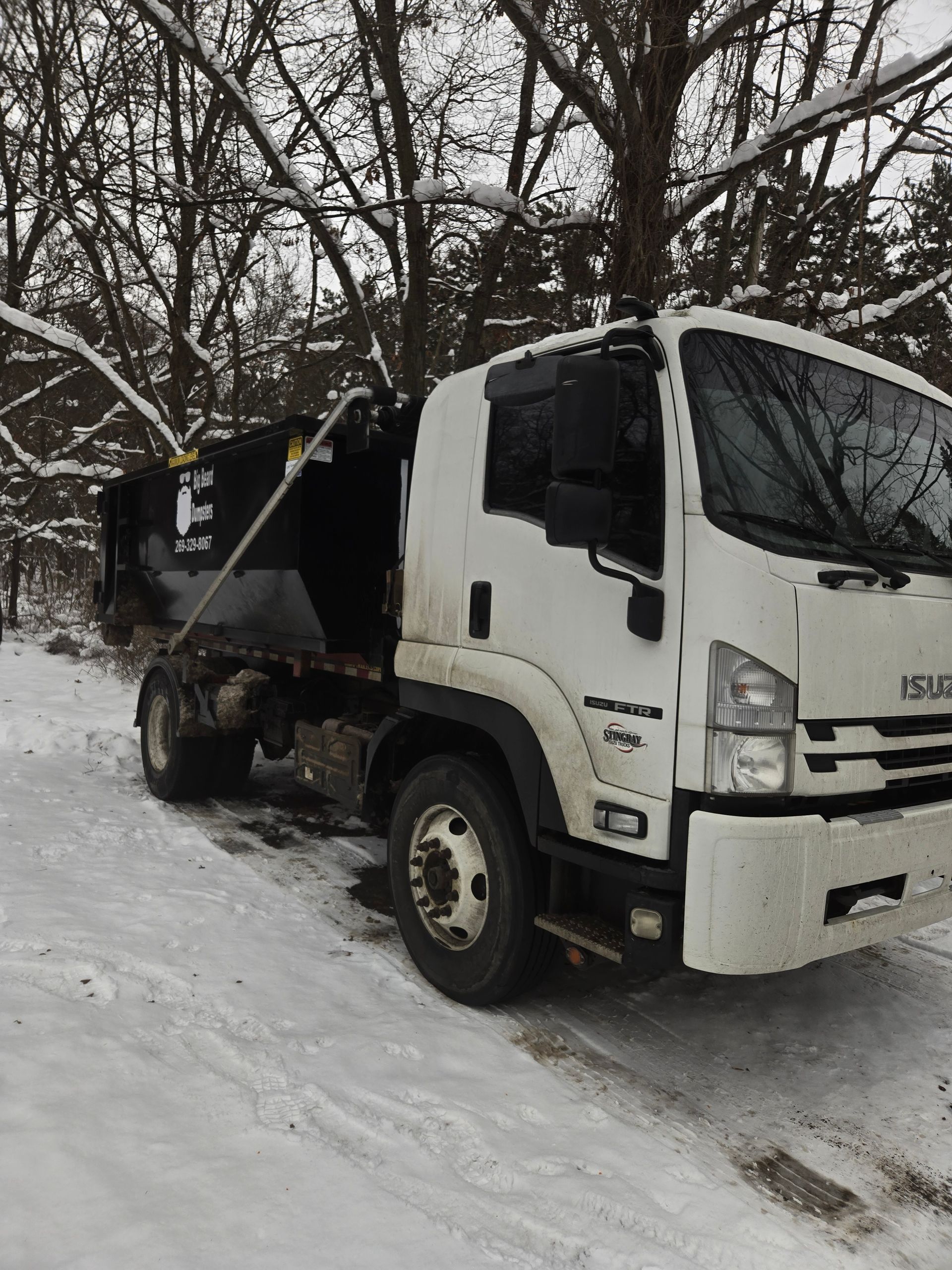 White Isuzu dump truck parked on snow-covered ground near bare trees.