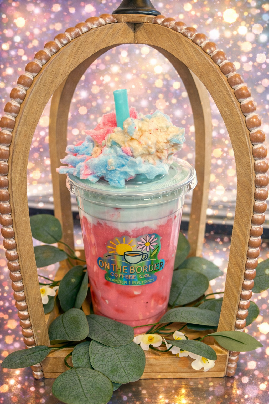 Cotton candy-topped drink in a clear cup with pink and blue layers, framed by wood and flowers.