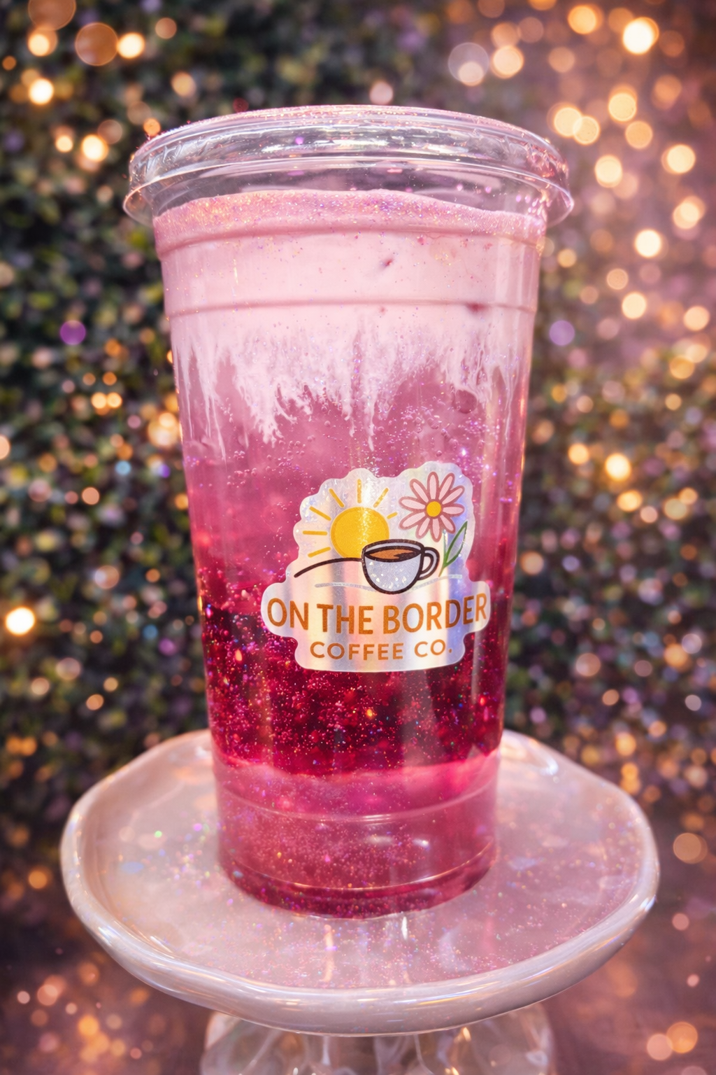 Pink glittery drink in a clear cup, with a coffee shop logo on a light-colored pedestal.
