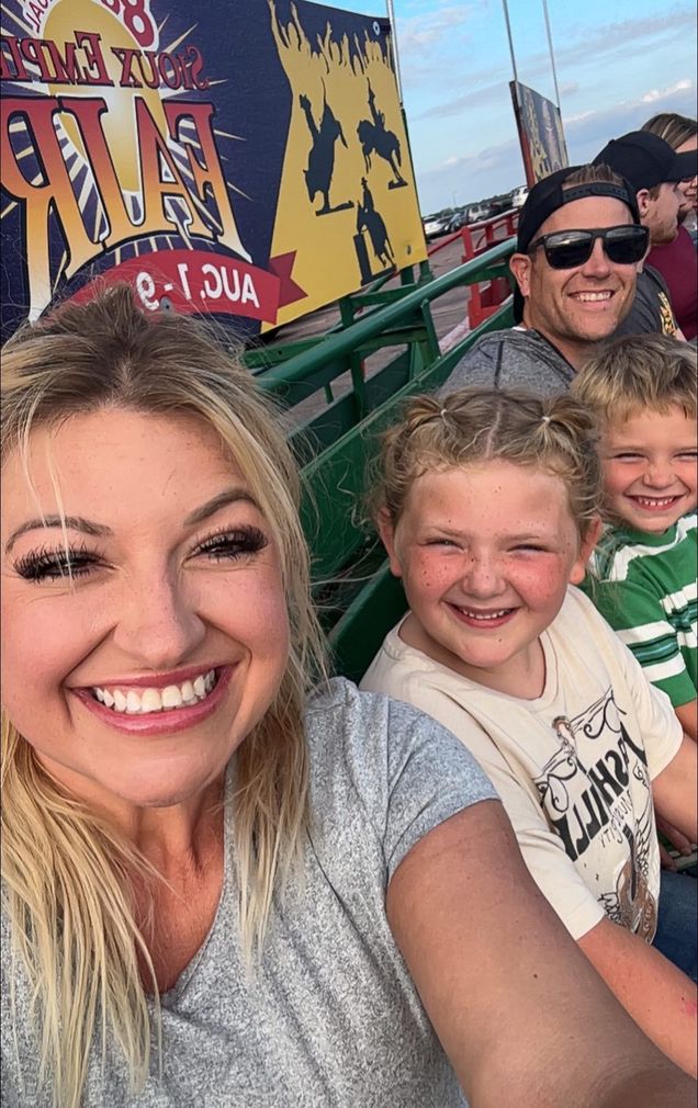 Family on a ride at an amusement park; smiling. Blonde woman takes selfie with kids and man in the background.