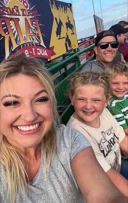 Family on a ride at an amusement park; smiling. Blonde woman takes selfie with kids and man in the background.