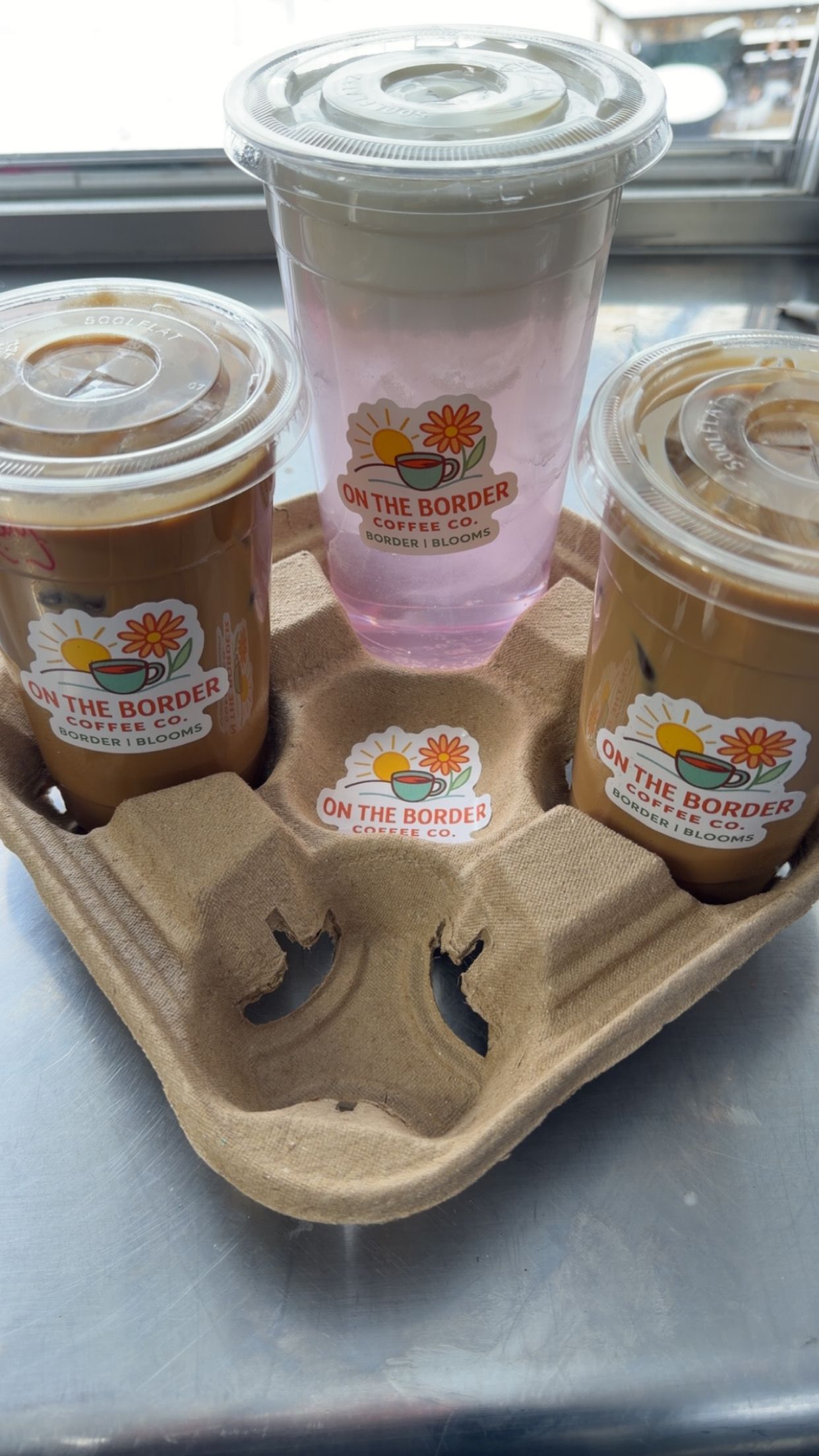 Three drinks in a cardboard carrier: two iced coffees and a pink drink, on a metal surface.