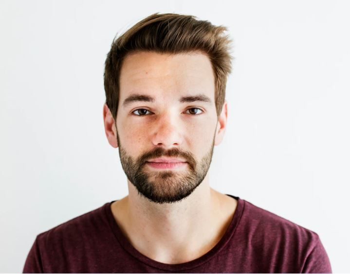 Hombre de cabello y barba castaños, vestido con una camisa color burdeos, mira a la cámara con una expresión neutra sobre un fondo blanco.