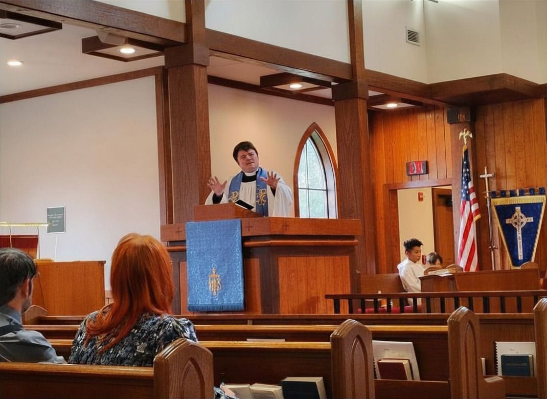 Fr. William dressed in white and blue stands in the pulpit of St. James giving a sermon with churchgoers sitting in the pews
