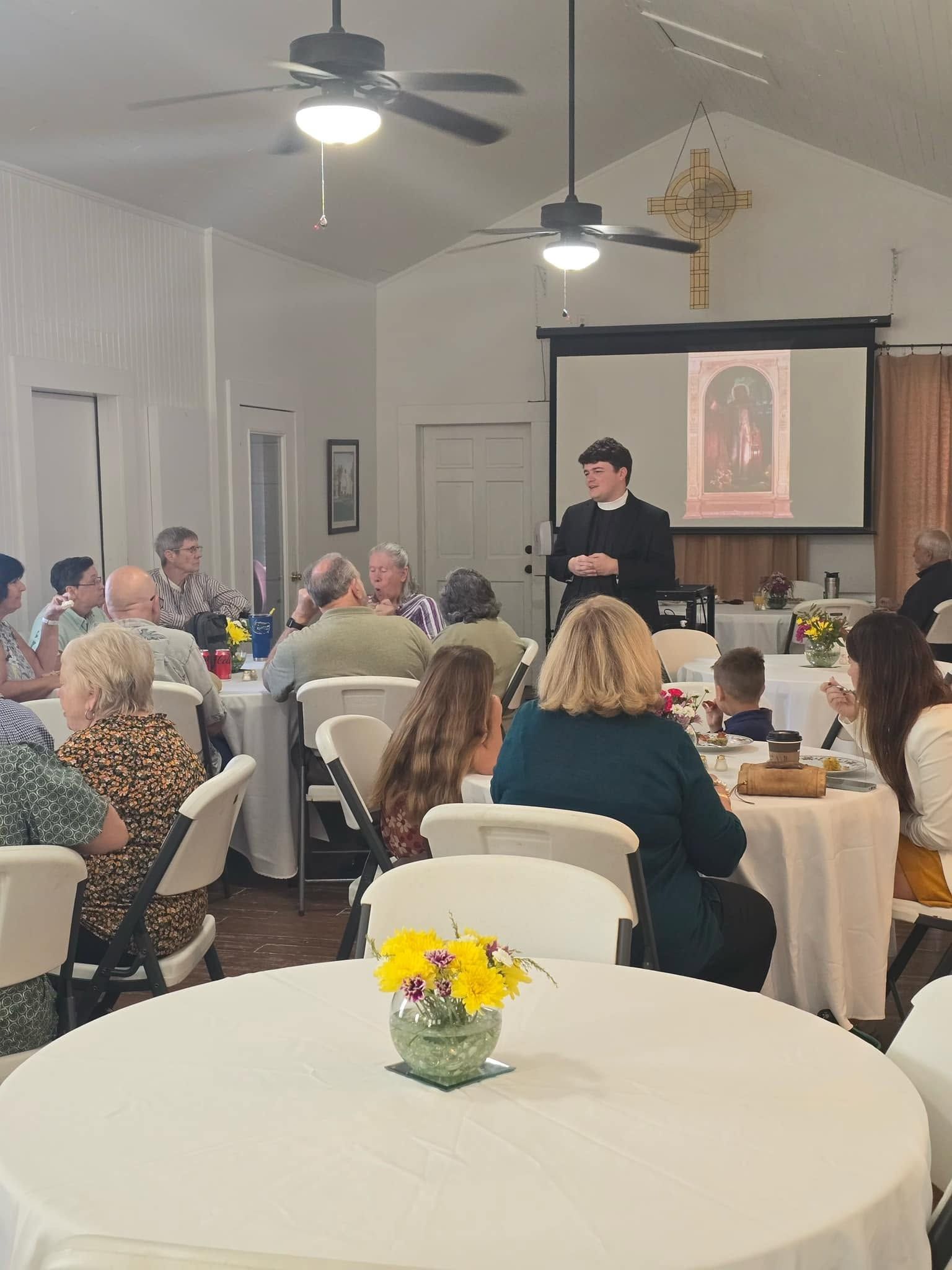 Fr. William stands giving a lecture to church members at Parish Hall in front of a projector screen