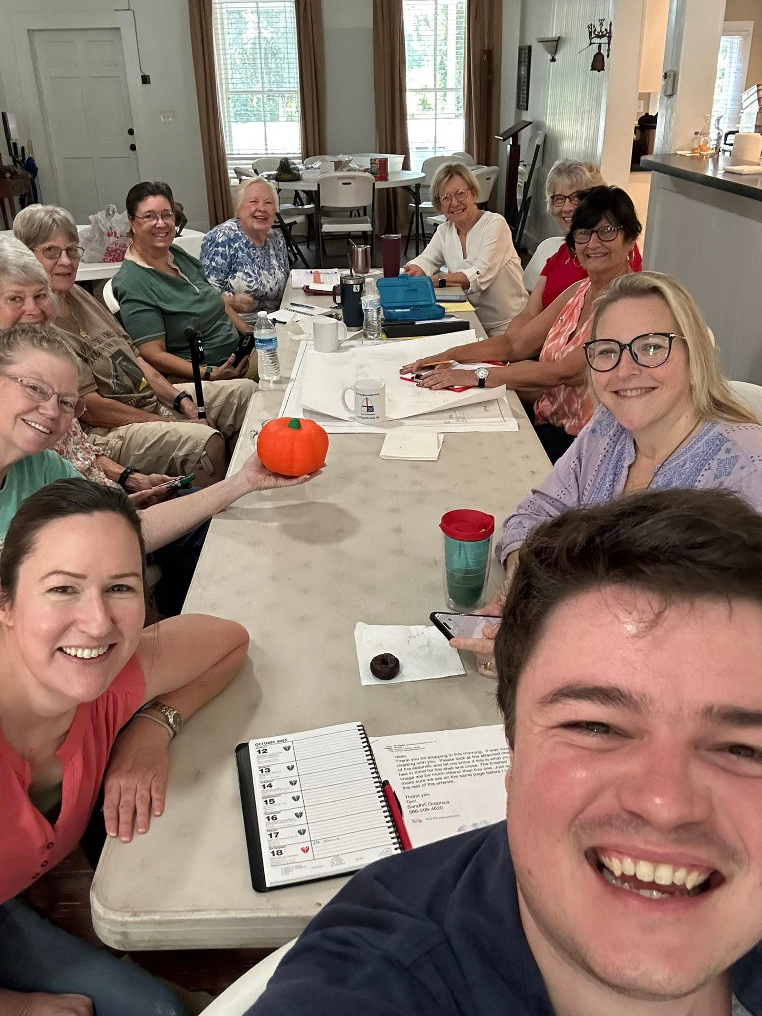 A group of church members are seated at a long table smiling with Fr. William taking a photo in front of them at Parish Hall