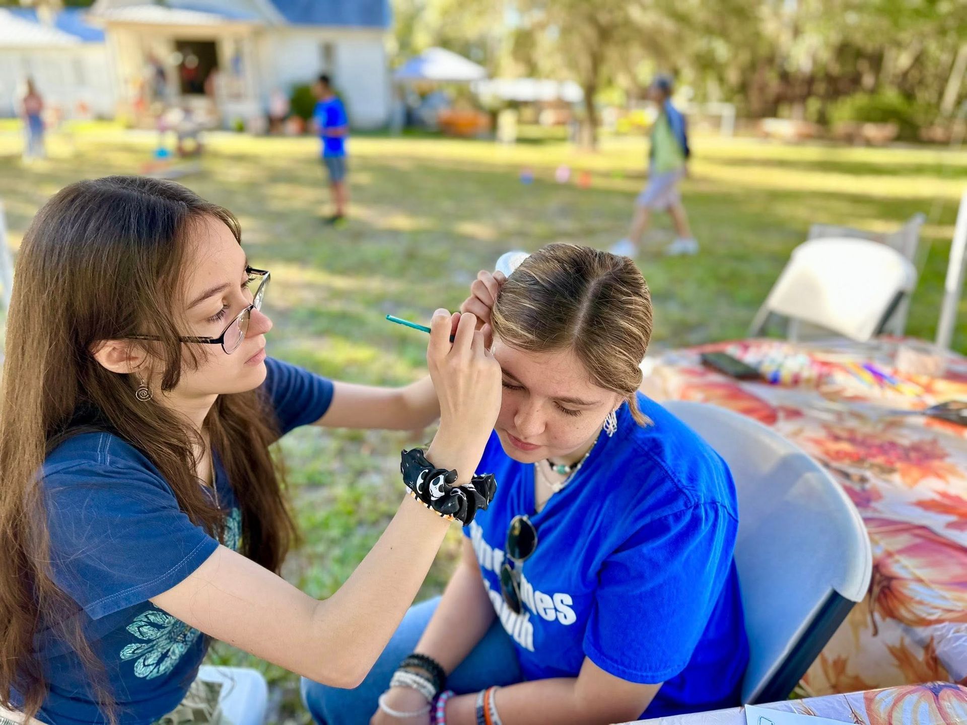 A youth member paints another youth member's face at Family Fall Festival with Parish Hall in the background