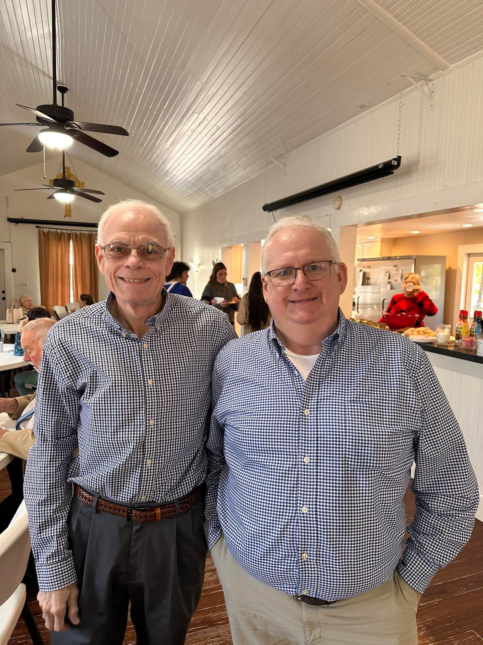 Two men pose for a photo with other members in the background inside St. James Parish Hall
