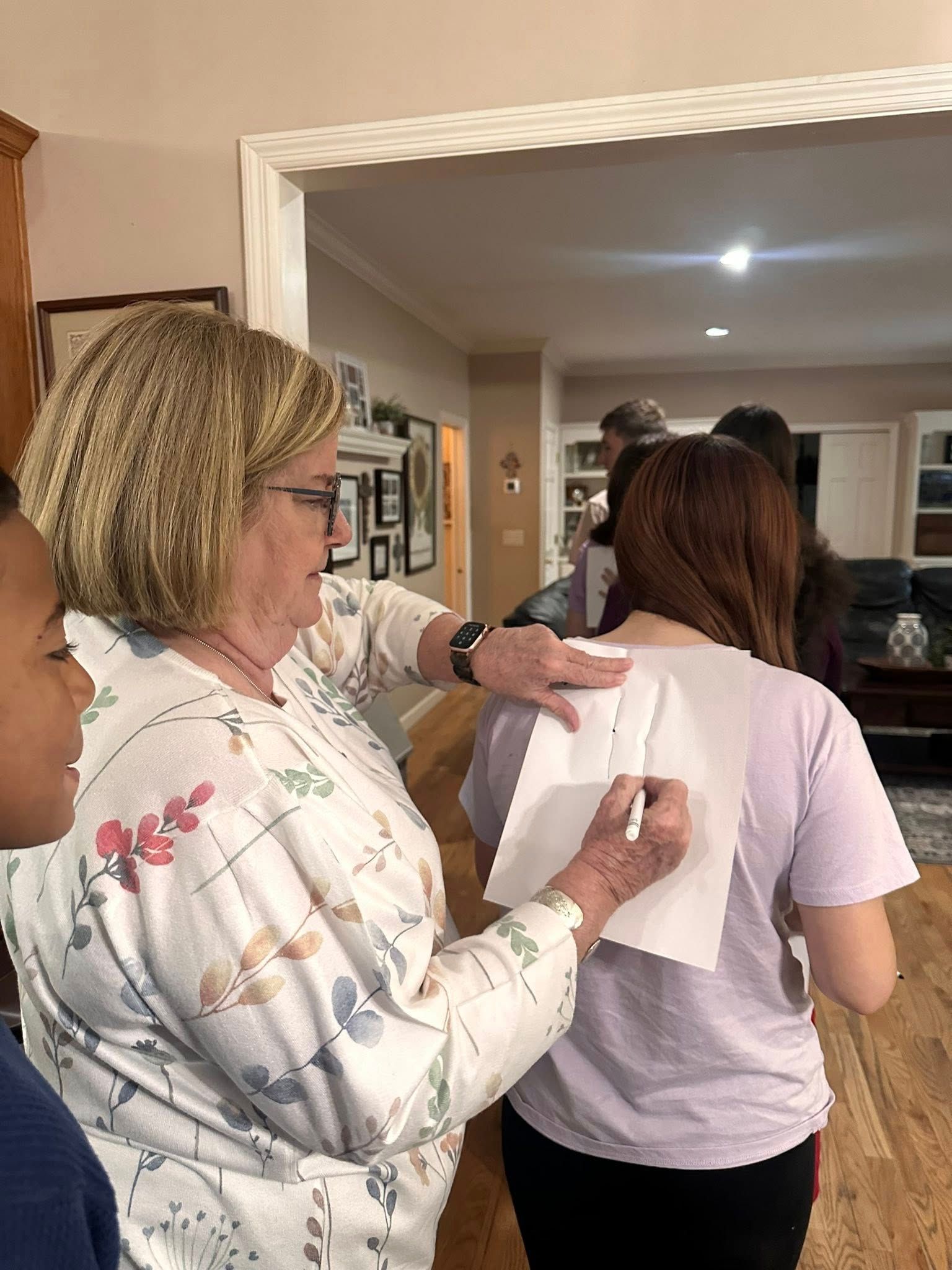 A woman is using a pen and paper on the back of another girl in an interior of a home