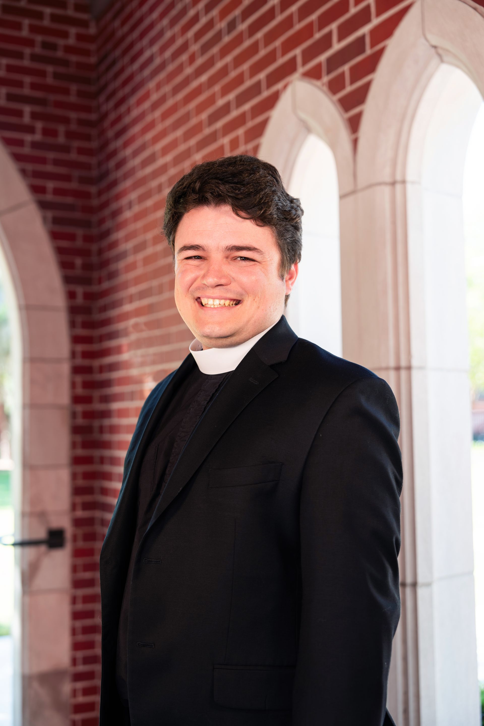 A picture of Fr. William smiling in front of an archway and brick exterior