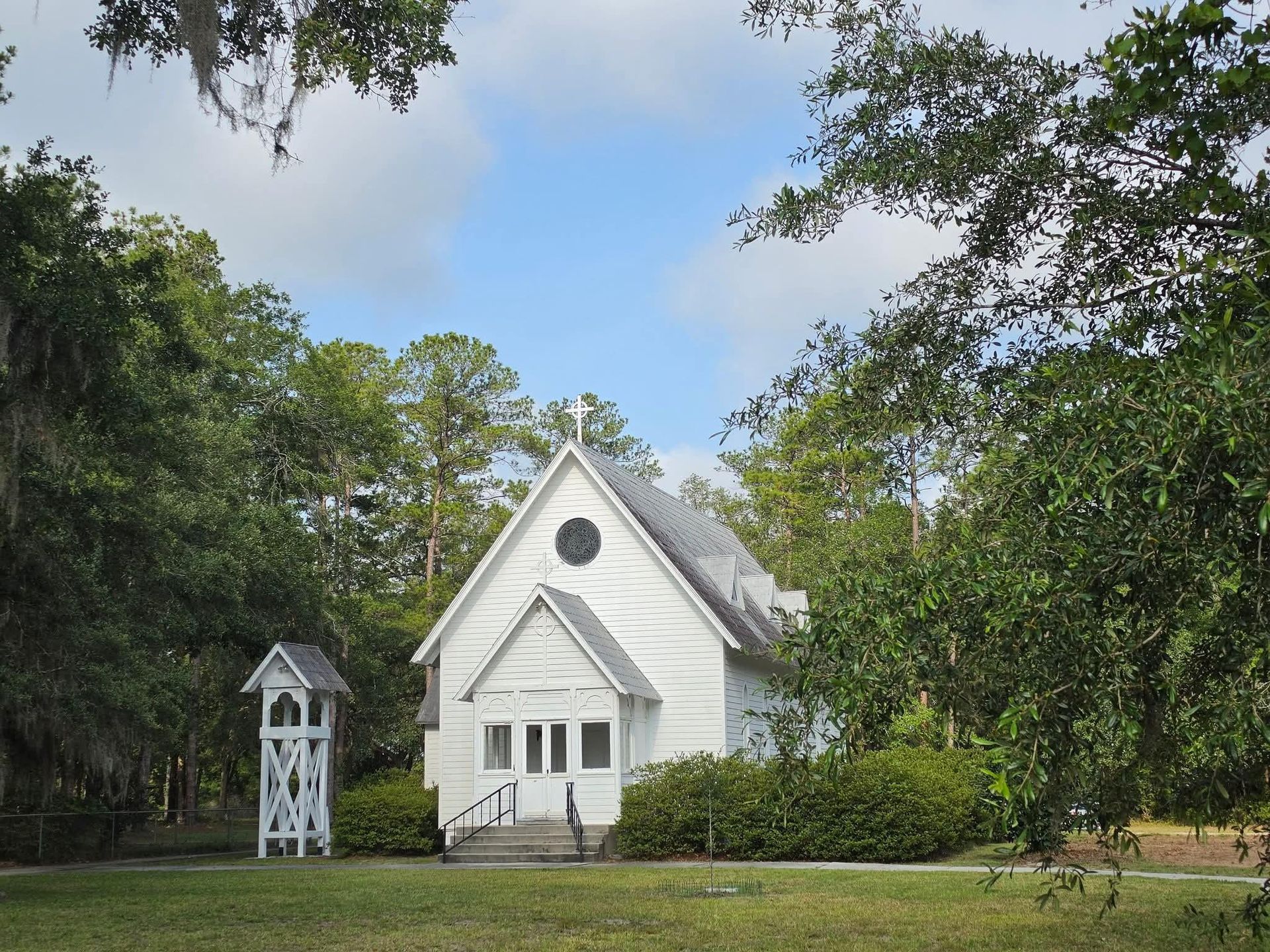 A white chapel nestled in between trees with blue skies above and a bell tower beside it