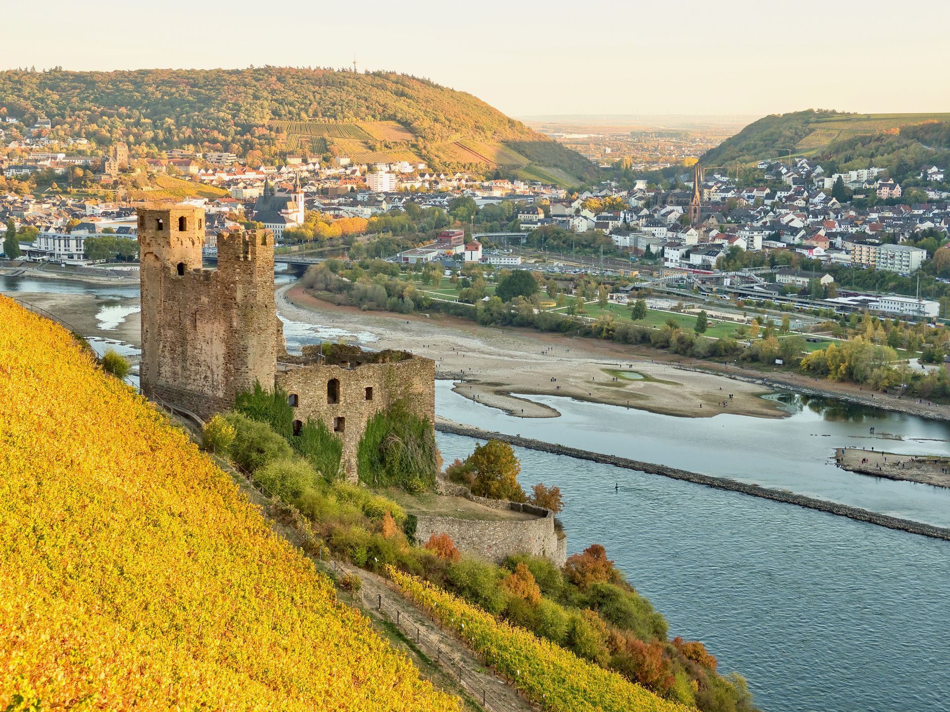 Stone castle ruins overlook a river and town in a sunlit valley with autumn hills.