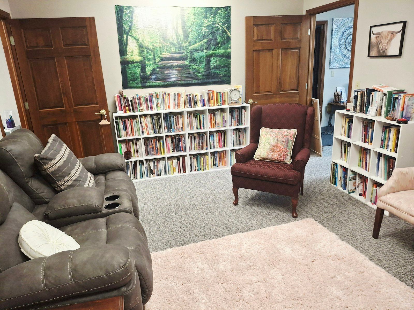 Cozy office with couch, chair, and bookshelves. A forest scene tapestry hangs on the wall.