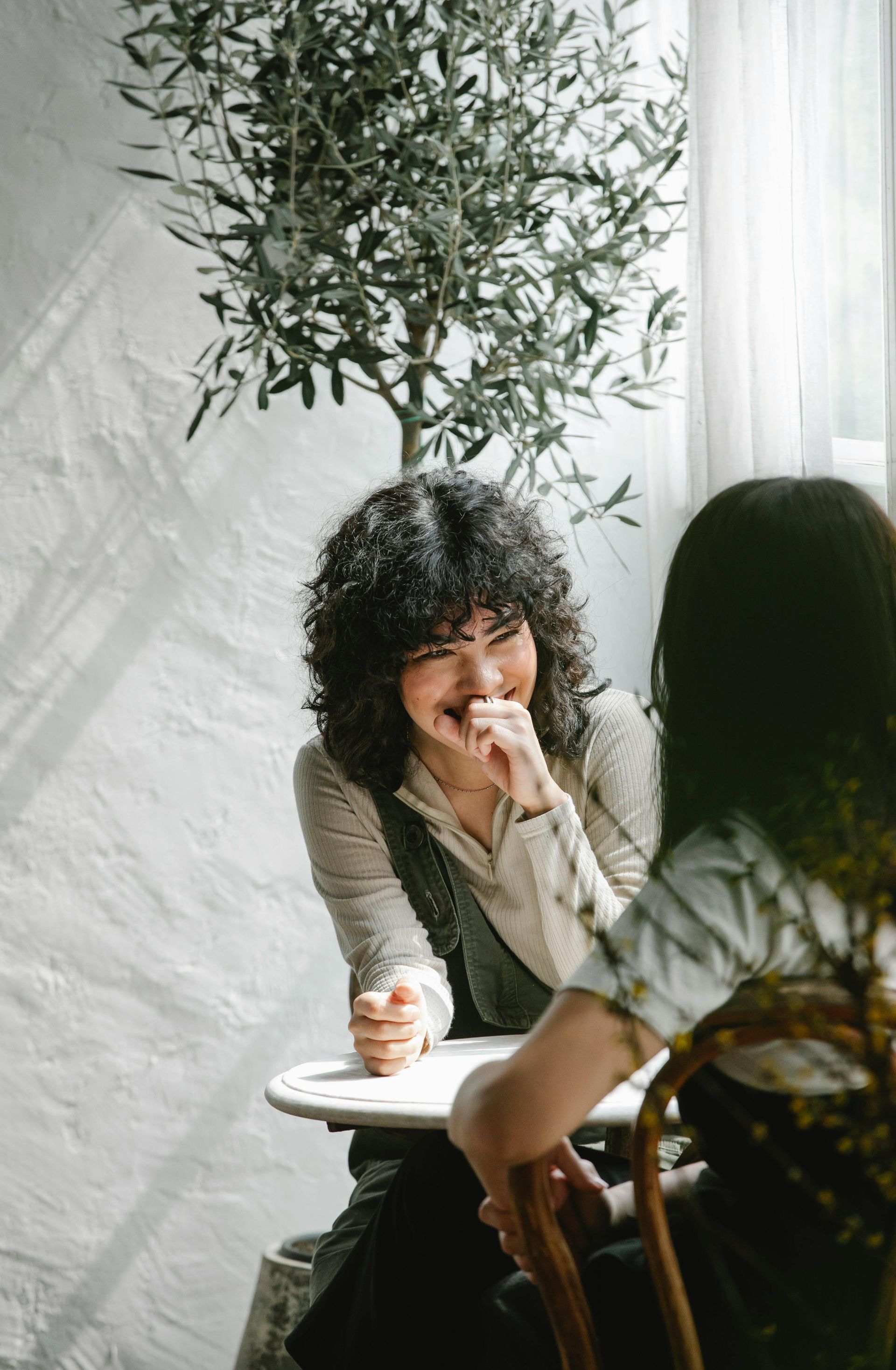 Two women chatting at a café table by a bright window, with a tall potted tree behind them.