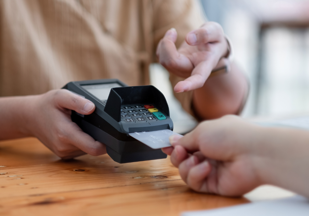 A person holding a payment terminal as another person inserts a card to complete a transaction on a wooden counter.