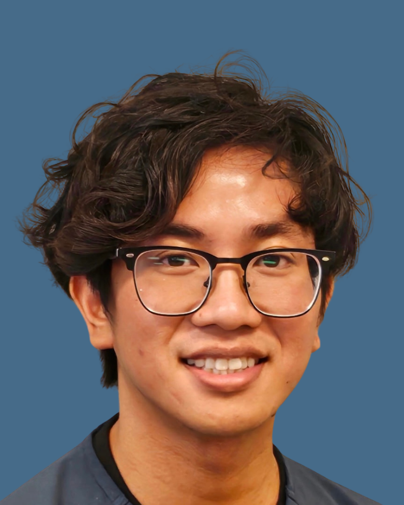 A person with wavy dark hair and glasses smiles warmly against a plain blue background.