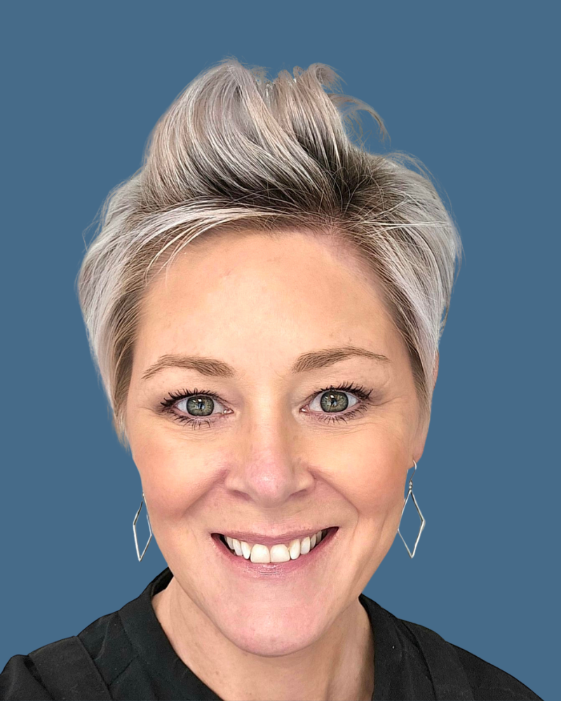 Headshot of a person with short, light-colored hair, smiling, wearing diamond-shaped earrings against a blue background.