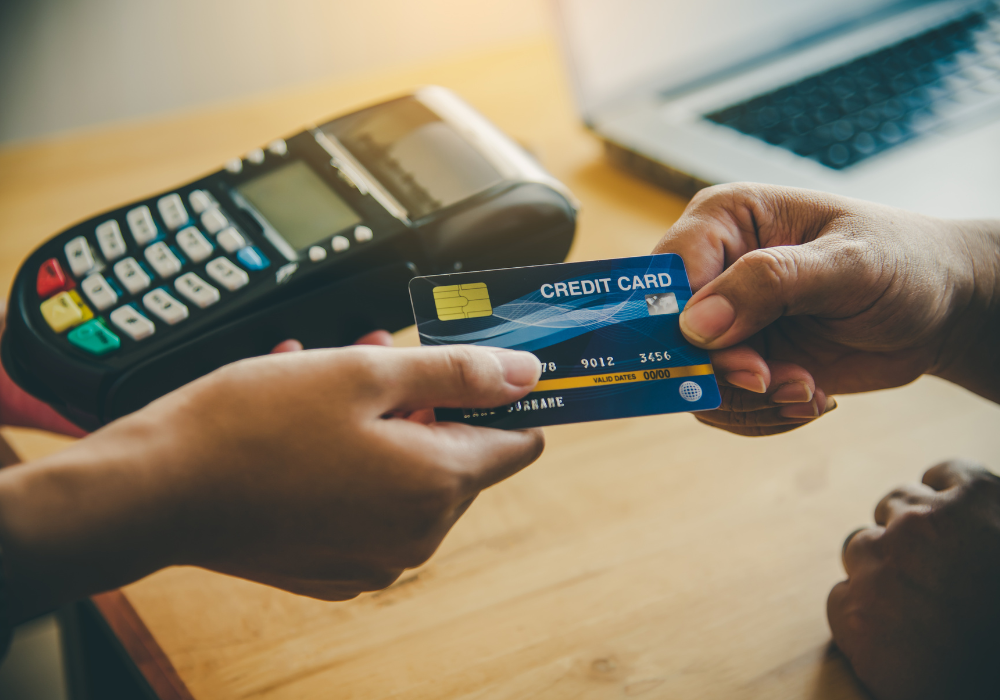 A person hands a blue credit card to another person near a payment terminal on a desk.