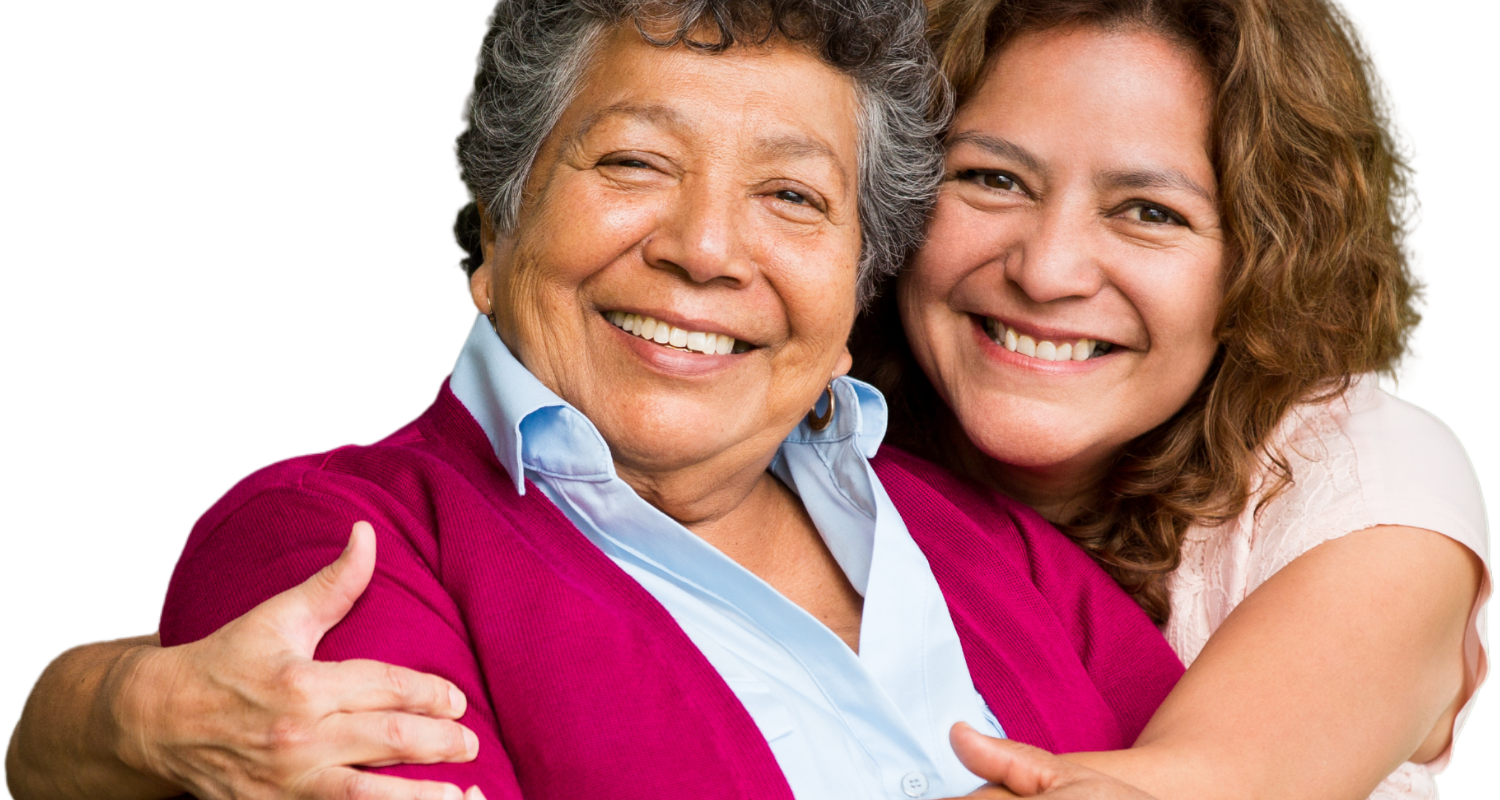 Two women smiling warmly, with one standing behind the other and embracing her, against a plain white background.