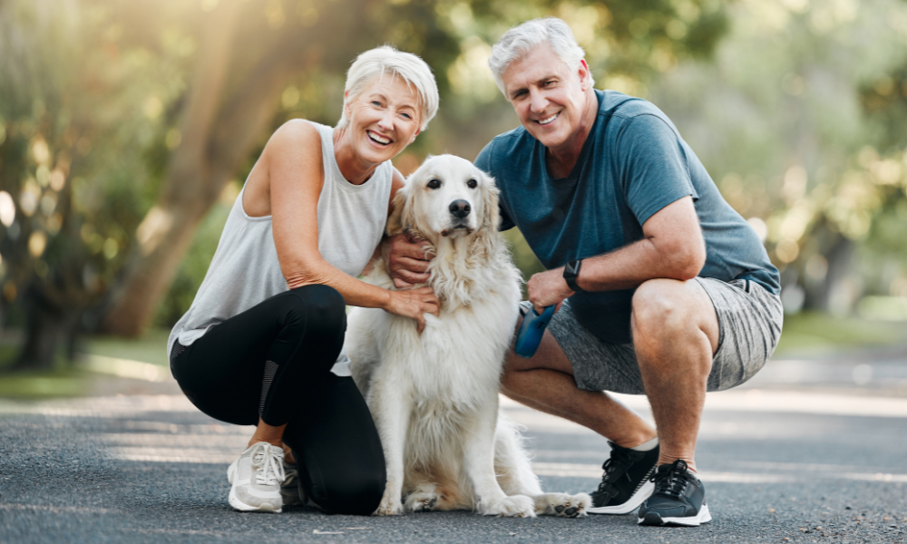 A smiling couple kneels on a path in a park, hugging their large, light-colored dog.
