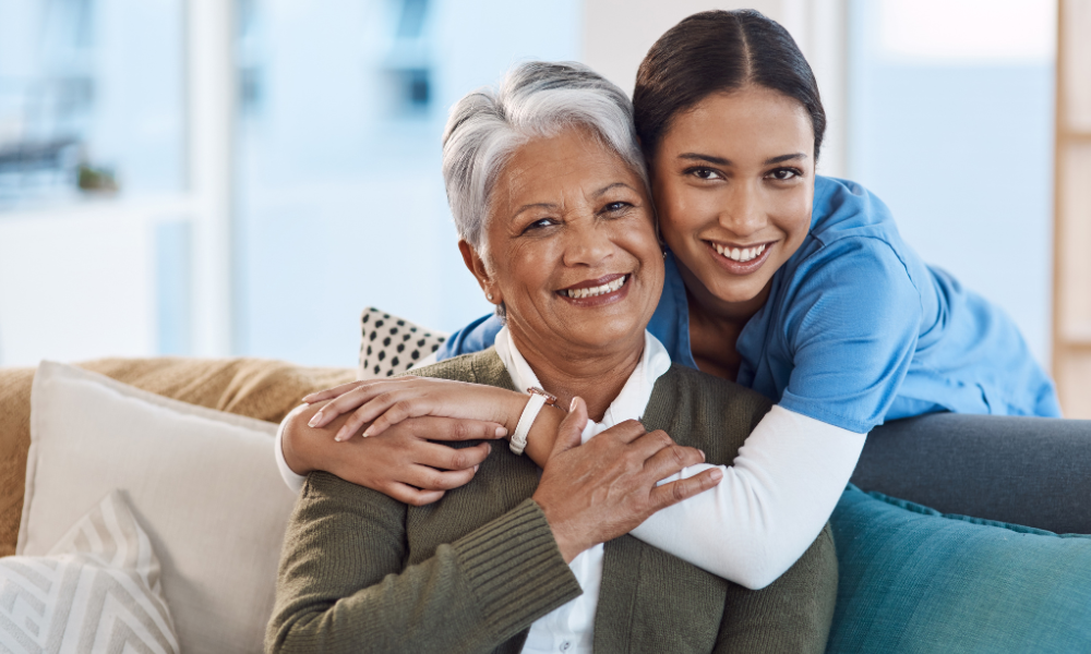 A caregiver in blue scrubs smiles while embracing a woman in an olive cardigan, both relaxing on a couch.