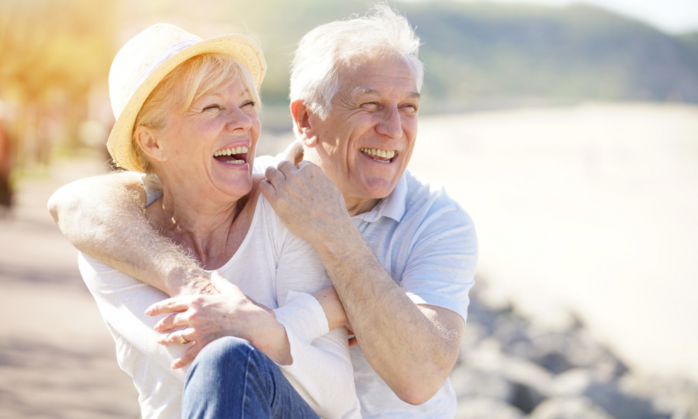 A smiling couple embraces outdoors near a beach, wearing light clothing and a hat, with a soft, sunny background.