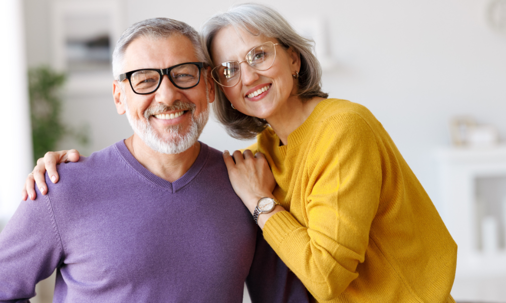 A smiling couple with gray hair and glasses posing together in an indoor setting.