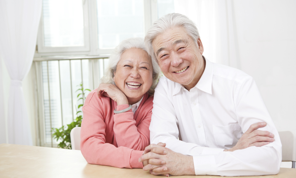 An older couple smiling broadly while sitting together at a wooden table in a bright, sunlit room.