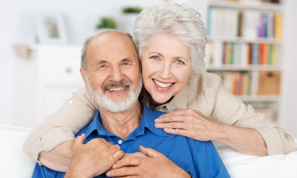 A smiling couple embraces while sitting together in a room with a bookshelf in the background.