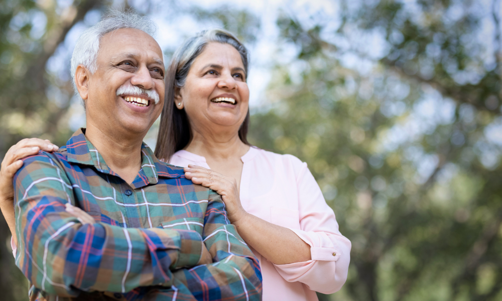 An older couple smiling, standing outdoors with a soft-focus background of trees.
