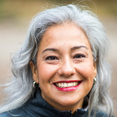 A person with silver-grey hair and bright red lipstick smiles warmly at the camera in a head-and-shoulders portrait.