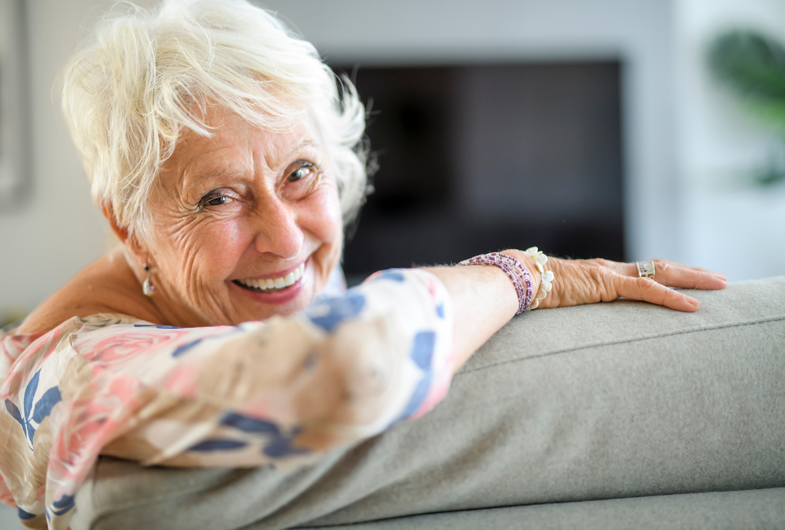 A smiling person with white hair, wearing a patterned top, resting their arms on the back of a couch.