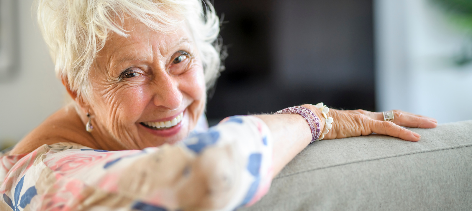 A person with short white hair smiles warmly while leaning back against a sofa, wearing a floral patterned top.
