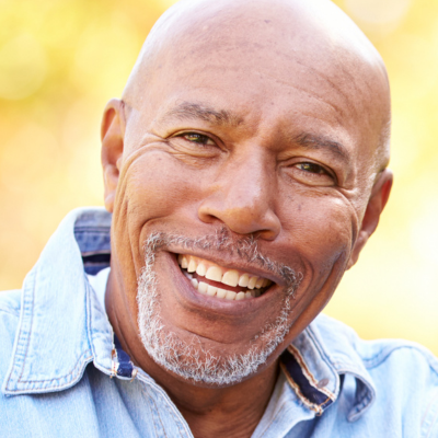 A smiling person with a shaved head and graying facial hair, wearing a denim shirt against a blurred, golden background.