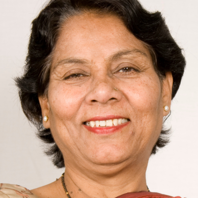 A person with dark, shoulder-length hair and small earrings smiles warmly against a plain, light-colored background.