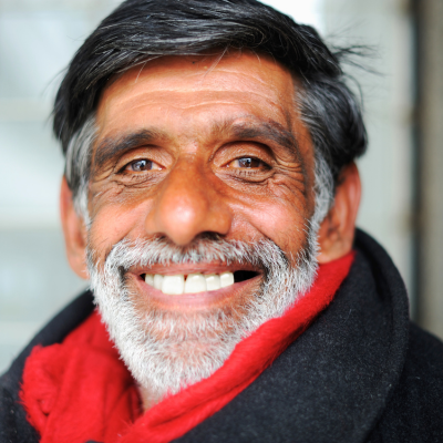 A close-up portrait of a smiling person with dark hair, a grey beard, and a bright red scarf against a blurred background.