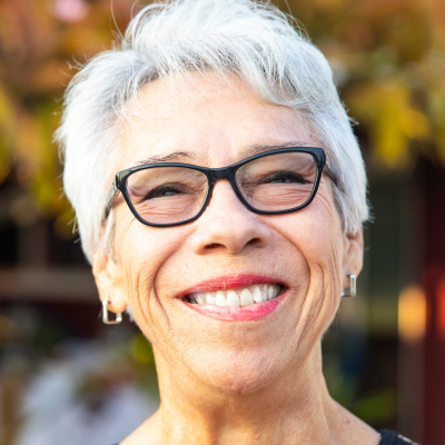 Smiling person with short grey hair and black-framed glasses, set against an out-of-focus outdoor background.