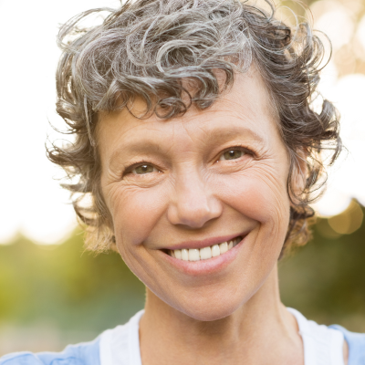 A person with curly grey hair smiling warmly at the camera outdoors with a soft, blurred natural background.