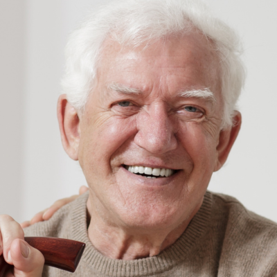 A smiling person wearing a beige sweater with one hand resting on a wooden cane.