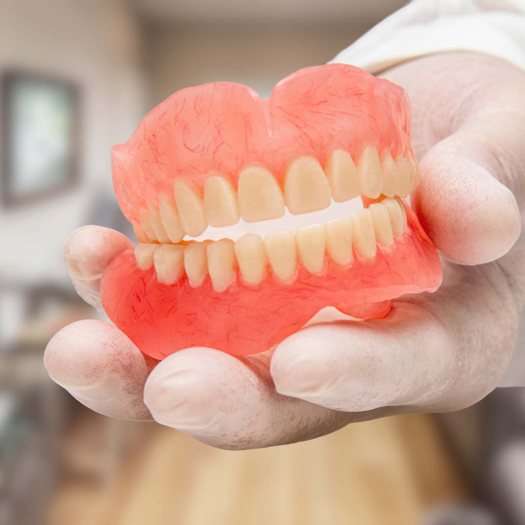 A gloved hand holds a complete set of upper and lower dentures against a blurred indoor background.