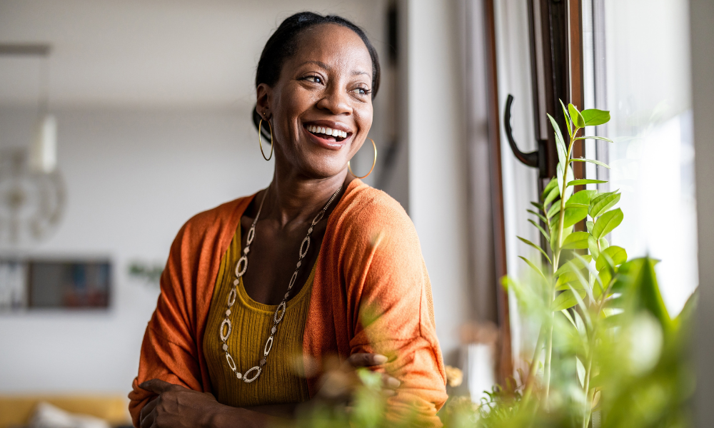 A smiling person wearing an orange cardigan and a long necklace, looking out a window near a green plant.