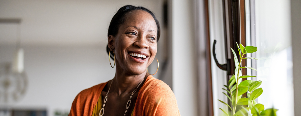 A person wearing an orange top smiles while looking out a window next to a green houseplant.