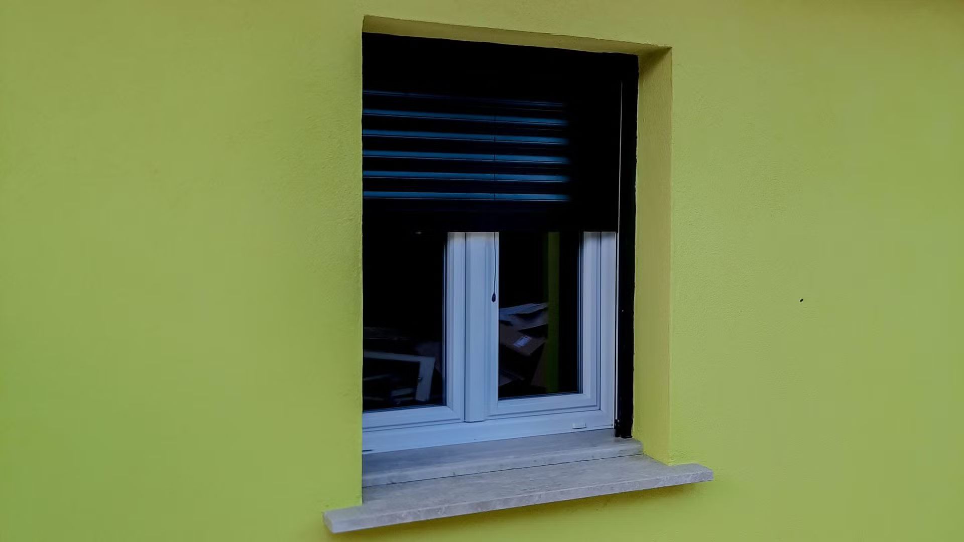 Un muro giallo con una finestra con una persiana nera.
