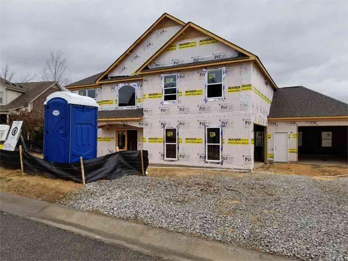 House under construction with blue porta-potty in front. Gravel and black tarp on the ground. Overcast day.