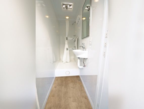 Bathroom interior with a shower, sink, and mirror. White walls, wood-look floor, and bright lighting.