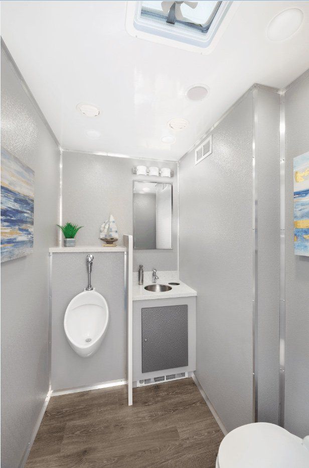 Interior of a luxury portable restroom with a urinal, sink, and toilet, all in neutral tones.